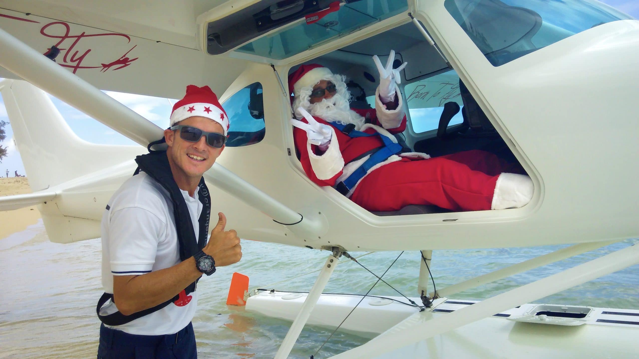 Une personne coiffée d'un bonnet de Père Noël lève le pouce à l'extérieur d'un hydravion, tandis que le Père Noël fait des signes de paix à l'intérieur.