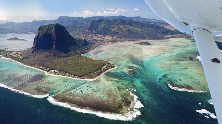 Aerial view of coastal landscape with a mountainous island, lush greenery, turquoise water, and a plane's wing.