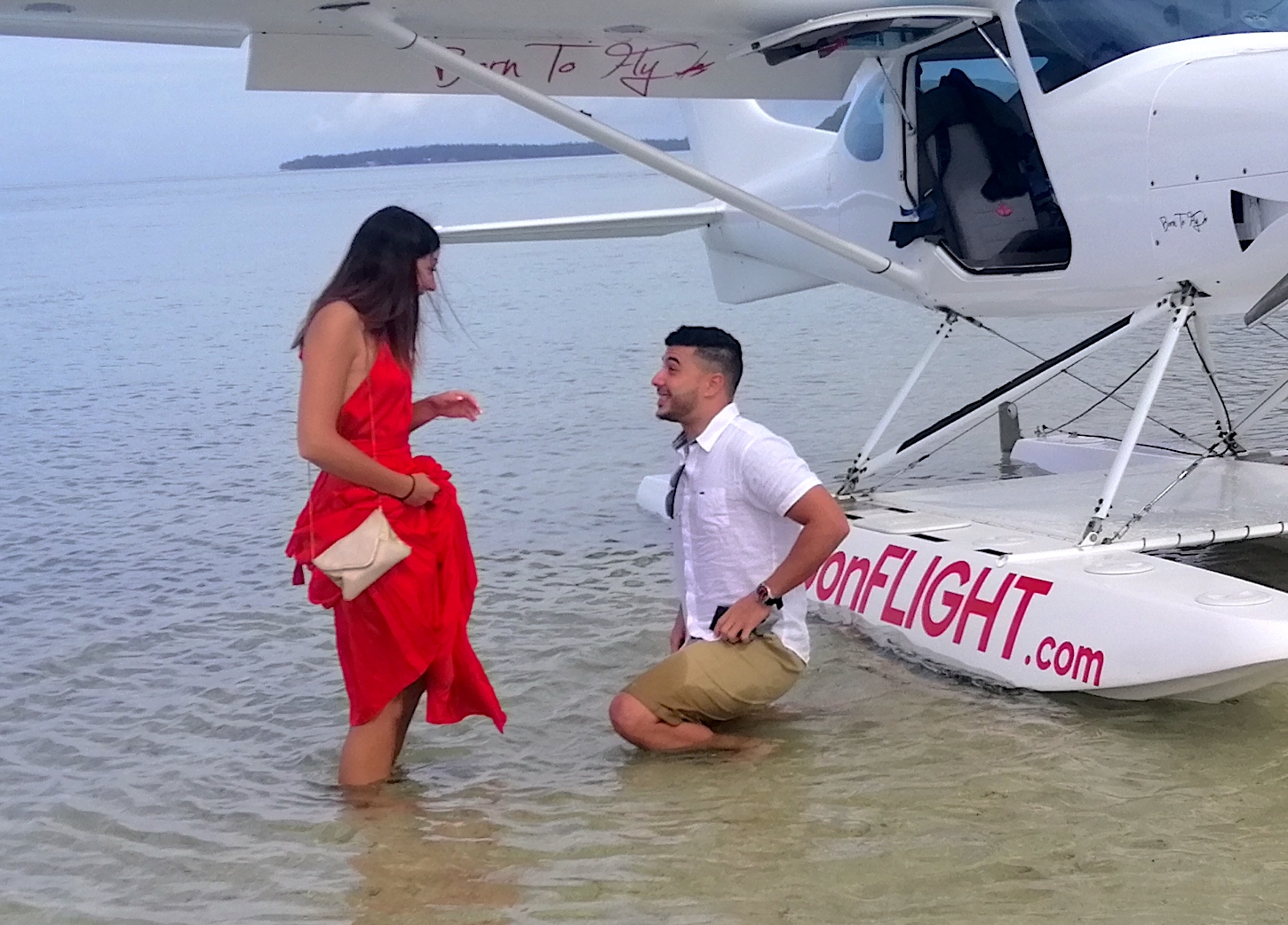 Man kneeling in water near woman by branded onFLIGHT.com seaplane at shoreline.